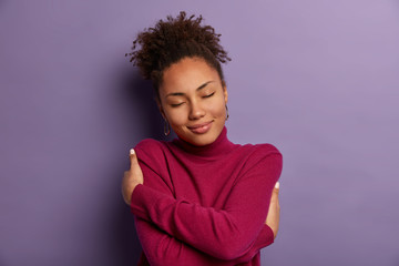 I love myself. Tender romantic woman embraces own body, hugs herself, closes eyes from pleasure, wears soft turtleneck just for cold weather, feels comfort, stands indoor against purple wall