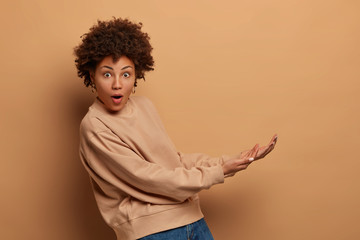 Isolated shot of surprised young Afro American woman pretends catching something big, keeps palms,...