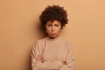 Fototapeta na wymiar Photo of troubled sad woman stands upset and offended, keeps arms folded, looks with regret from under forehead, wears casual beige jumper, expresses negative emotions, blows cheeks from anger