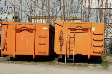two roll on/off containers in orange color on one of them is leaned a metal ladder to facilitate access. They are big volume construction waste containers.