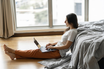 Beautiful young brunette girl working on a laptop, sitting on the floor near the bed by the panoramic window with a beautiful view from the high floor. Stylish modern interior. A cozy workplace