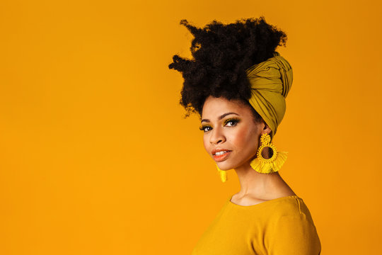 Portrait of a serious young woman with big yellow tassel beaded earrings and afro hair wrapped with head wrap scarf, isolated on yellow background