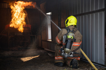 Obraz premium Rear view of Fireman wear fire protection suit walking into the fire training room and Spray water Fire at fire station. Training fire drill Concept.