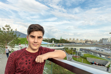 A teenager with dark hair and brown eyes is leaning on a railing against a blue sky with white clouds.