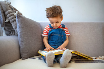 Little, cheerful, three years old boy on the sofa, with open book in front of him. Cute little boy...