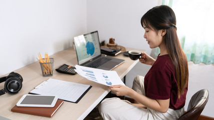 Attractive young beautiful asian woman working with laptop and document while sitting at the indoors living room office as a freelancer, e-coaching working, remotely or work from home concept.