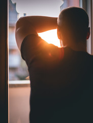Young man looking through the window of his home during a coronavirus quarantine at sunset