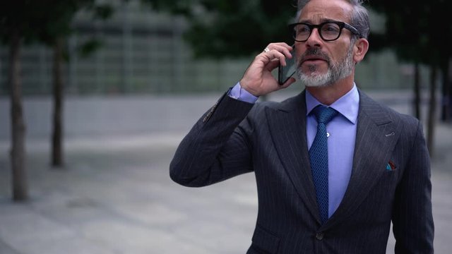 Mature entrepreneur in optical eyewear making international conversation with business partner connected to roaming internet on modern smartphone gadget during time in downtown
