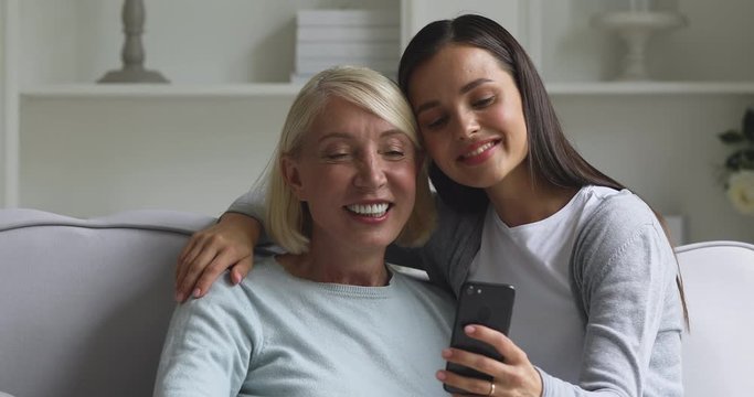 Smiling attractive young lady embracing happy sincere older senior mommy, posing for selfie photo on smartphone. Affectionate two generations family watching funny videos on smartphone.