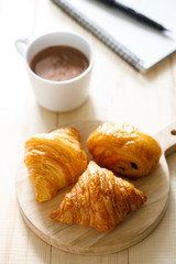 A cup of hot chocolate and croissant on wooden background with notebook.  Work from home.