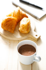 A cup of hot chocolate and croissant on wooden background with notebook.  Work from home.
