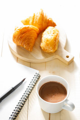 A cup of hot chocolate and croissant on wooden background with notebook.  Work from home.