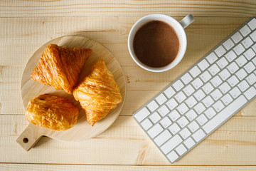 A cup of hot chocolate and croissant on wooden background with computer keyboard.  Work from home.
