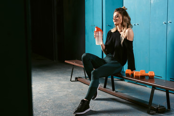 Ready to workout in gym! Beautiful woman in sportswear is holding a shaker and sitting in locker...