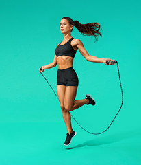 Sporty girl exercising with skipping rope. Photo of girl in black sportswear on turquoise background. Dynamic movement. Full length. Sports and healthy lifestyle - obrazy, fototapety, plakaty
