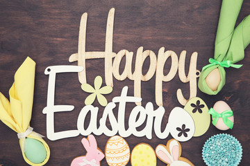 Text Happy Easter with eggs and gingerbread cookies on brown wooden table