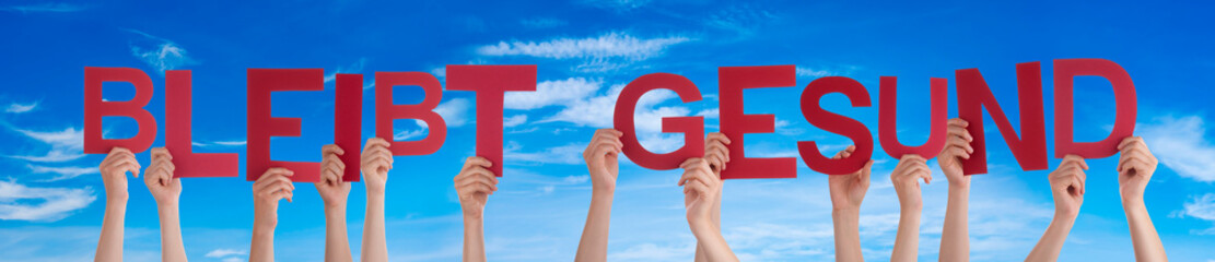 People Hands Holding Colorful English German Bleibt Gesund Means Stay Healthy. Blue Sky As Background