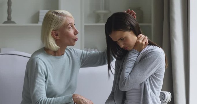 Compassionate caring older mature woman stroking grownup stressed upset daughter, sitting together on sofa. Empathic middle aged granny supporting soothing unhappy young granddaughter at home.