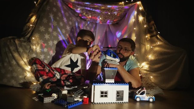 Father and son have fun playing with plastic constructor blocks while sitting in a makeshift blanket tent indoors. Father with little son have fun at home in quarantine.