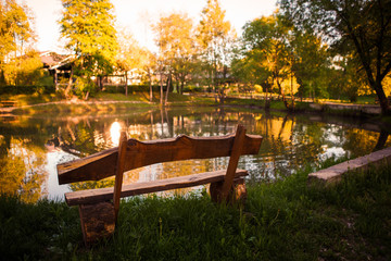Fototapeta na wymiar Wooden bench on the Bank of a pond in the Park. A picturesque place to relax in nature in the orange light of the sunset