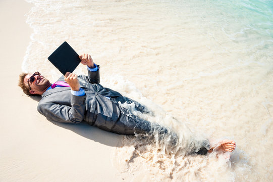 Barefoot businessman relaxing with a tablet computer on the shore of a tropical beach with waves breaking over his suit