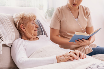 Helpful young carer reads a book to an elderly woman lying in a hospital bed