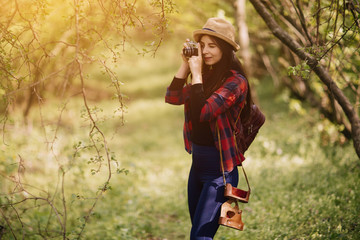 stylish hipster young woman  taking photos in the forest, carrying a backpack in the forest on sunset light in the spring season, looking at amazing woods, travel concept
