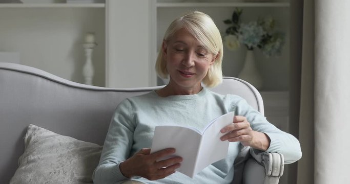 Happy mature elderly woman resting on couch, involved in favorite paper book reading at home. Pleasant middle aged smiling retired granny enjoying spending hobby leisure time alone in living room.