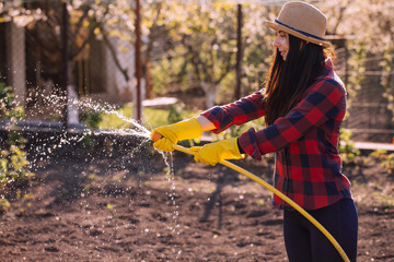 young woman in a straw hat and a red plaid shirt holds a yellow hose watering the garden,On the background flowering spring trees. Organic farming and spring gardening concept