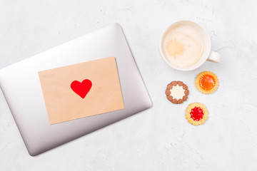 Top view of closed laptop with craft envelope and red heart, cup of coffee and come cookies. Desktop. Love care St. Valentine's Day Mother's Day concept. Free space. Copy space. 