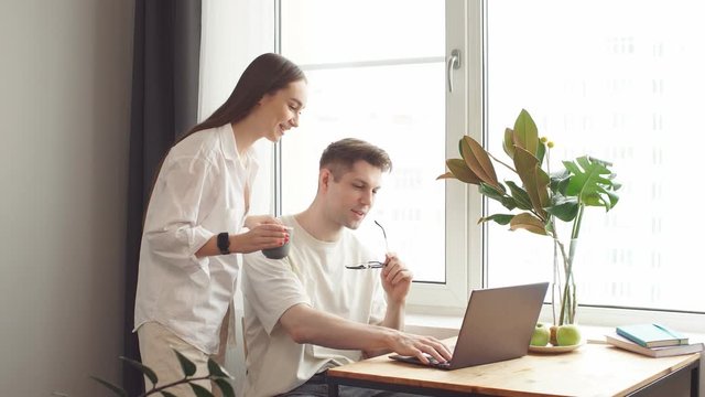 Pretty caucasian wife support her husband while he is working at home, man sit with laptop. Couple in domestic clothes.