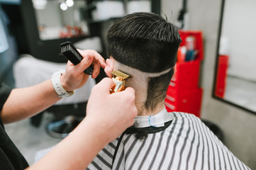 Professional barber creates a stylish hairstyle for the client with a golden clipper in his hands and a close-up photo. Hands of a barber with a golden trimmer cuts the hair of a brunette man
