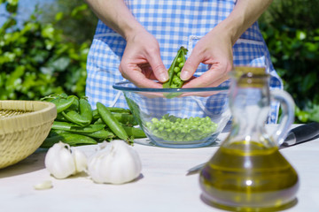 Close up of the hands of a woman shelling peas from the pod inside a glass bowl outdoors with sunlight in the greenery