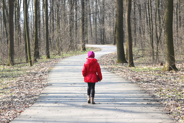 Little girl in red jacket and red hat walks in spring forest among trees on asphalt road