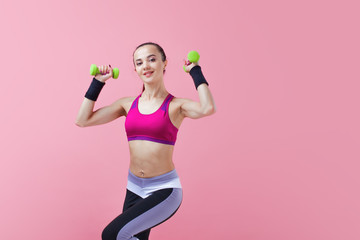 Young athletic woman, in a bright sports top, with green dumbbells, raises her arms and knees, on a pink background.