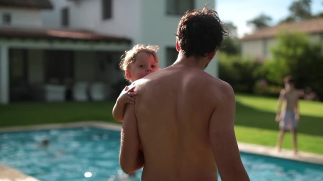 Back of father holding baby infant boy in arms at swimming pool outdoors. Dad bonding with toddler son