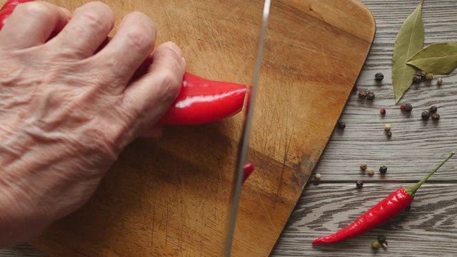 Using Knife To Cutting Ramiro Red Hot Pepper On Cutting Wooden Board. Top View. Close Up. Green Bay Leaf, Multi Color Peppercorns And Bright Red Chili Pepper On Gray Wooden Background..