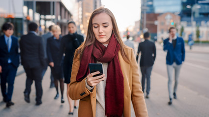 Young Smart Casually Dressed Female is Using a Smartphone on a Street. Business People and Office Managers Walk Pass on Their Way to Work. She Looks Confident while Checking Her Cell and Walking.