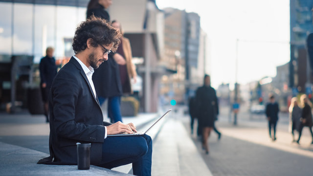 Handsome Businessman in a Suit is Sitting on Steps next to Business Center and Working on a Laptop on a Street in a City. Office People Walk By to Work.