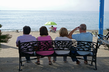Four people sitting on a bench on the promenade looking at the sea and a person taking a sun bath under a parasol