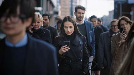 Fototapeta premium Beautiful Businesswoman in Black Coat is Using a Smartphone on a Street in Downtown. She Walks on a Crowded Pedestrian Street and Looks Successful. She's Browsing the Web on Her Device.