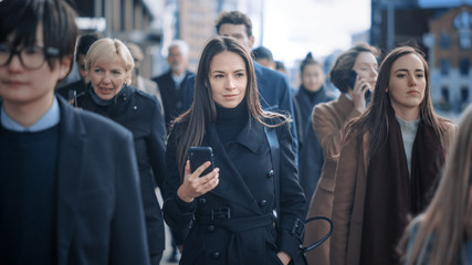 Beautiful Businesswoman in Black Coat is Using a Smartphone on a Street in Downtown. She Walks on a Crowded Pedestrian Street and Looks Successful. She's Browsing the Web on Her Device.