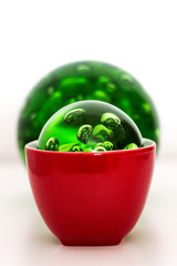 una esfera verde metida en una taza roja con otra esfera verde más grande detrás en fondo blanco