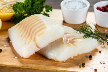 Fresh raw cod with seasonings and vegetables served on cutting board on wooden table
