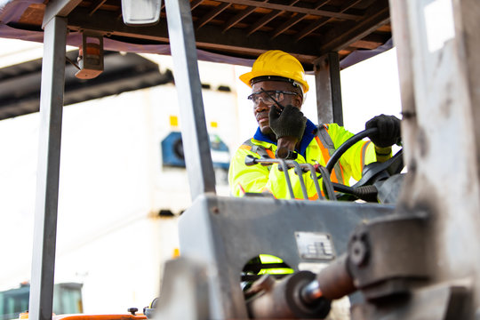 African American Man at work. Professional operation engineering. Young worker forklift driver wearing safety goggles and hard hat sitting in vehicle in warehouse. Walkie talkie communication