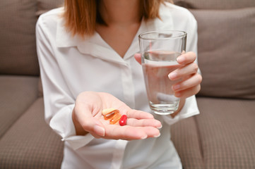 Close up Asian woman holding pill and a glass of water sitting on sofa. Concept of taking daily medicine, Multivitamins and supplements and medicine to cure head ache, stomach pain sedation meds.