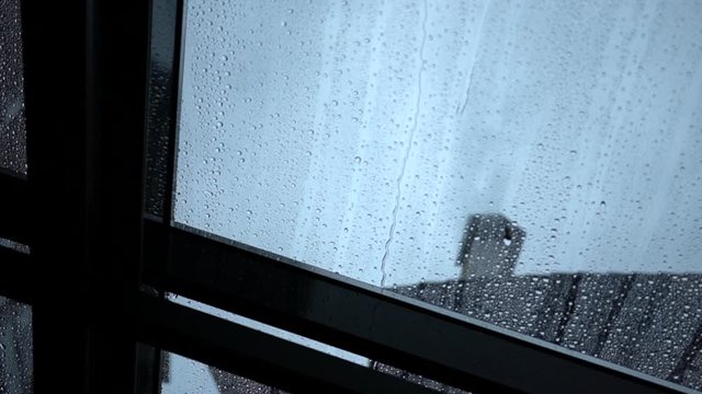 Rain drops slowly flow down the transparent roof window leaving water traces, close-up. A view through flat roof light on a dark building and grey sky background. Gloomy rainy day.