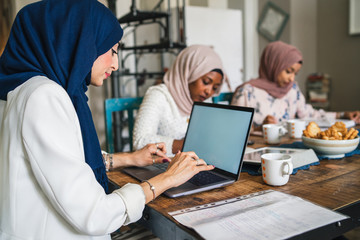 Obraz premium Three young Muslim women study on books and laptop at the kitchen table - Arabian millennials are preparing for university exams