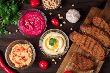 Plate of spicy traditional hummus with pepper and greens on a wooden background. Colorful hummus bowls decorated with chickpeas, herbs, garlic, tomate , crispy bread and chili pepper. Top view
