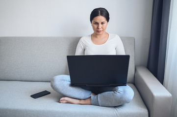 Young european freelance woman working on a laptop and phone on grey couch during coronavirus isolation home quarantine. Covid-19 pandemic Corona virus. Distance online work from home concept.